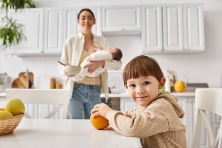 joyous mother in casual attire preparing breakfast for her toddler son while holding her newborn