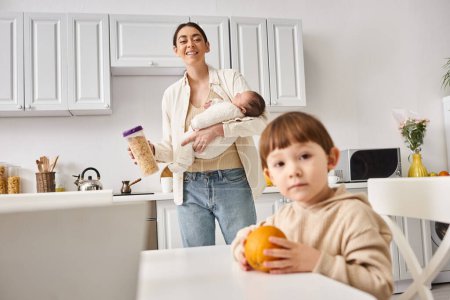 cheerful mother in casual attire holding cornflakes near her toddler son while holding her newborn
