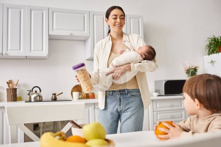 happy mother in casual attire holding cornflakes near her toddler son while holding her newborn