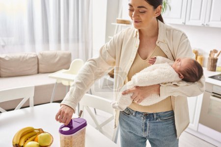 attractive happy woman in comfy homewear holding her cute newborn baby while preparing breakfast