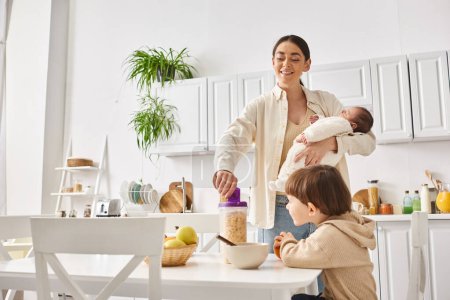 jolly beautiful woman pouring corn flakes to her adorable toddler son while holding her newborn baby
