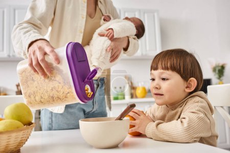 joyous woman pouring corn flakes to her adorable toddler son while holding her newborn baby