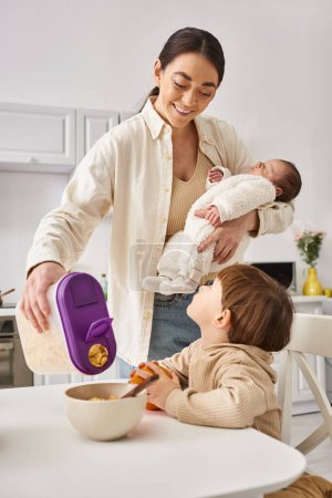 merry woman pouring delicious corn flakes to her adorable toddler son while holding her newborn baby