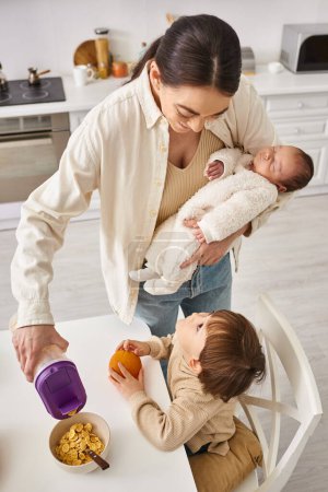 merry beautiful woman pouring corn flakes to her adorable toddler son while holding her newborn baby
