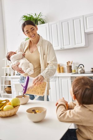 happy beautiful woman pouring corn flakes to her adorable toddler son while holding her newborn baby