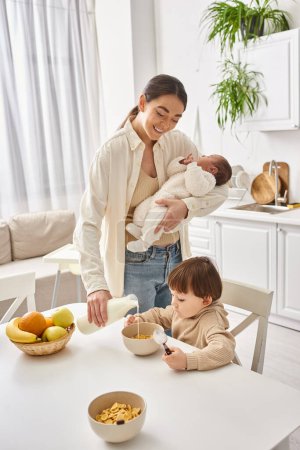fröhliche Frau gießt Milch in Cornflakes für ihren kleinen Sohn und hält ihr neugeborenes Baby