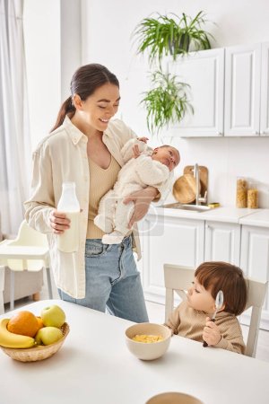cheerful beautiful mother enjoying breakfast with her toddler and newborn sons, modern parenting
