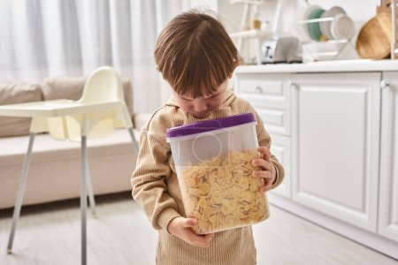 adorable toddler boy in casual homewear holding pack of corn flakes during breakfast on kitchen