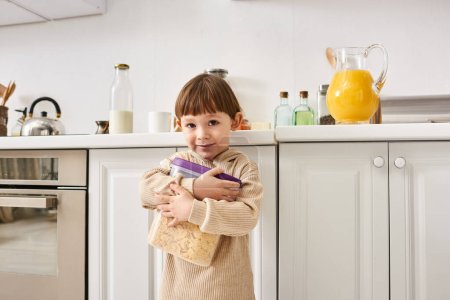 adorable little boy in comfortable homewear holding pack of corn flakes during breakfast on kitchen