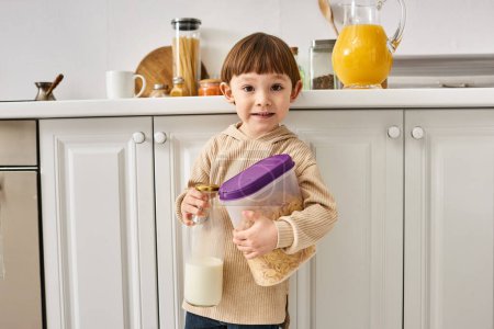 adorable toddler boy in cozy homewear holding milk and corn flakes during breakfast on kitchen