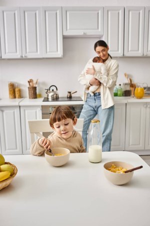 joyous loving mother posing next to her toddler son with newborn in hands during breakfast