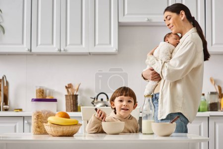 joyous attractive mother posing next to her toddler son with newborn in hands during breakfast