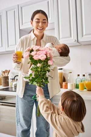 adorable toddler boy giving flowers to his attractive merry mother with his newborn brother in hands