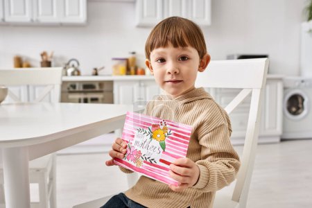 entzückender kleiner Junge in bequemer Kleidung sitzt am Muttertag und hält eine Grußkarte in der Hand