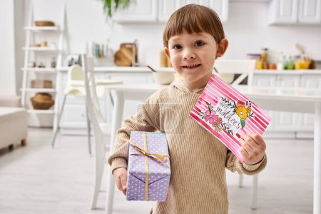 adorable lindo niño en cómoda ropa de casa posando con presente y tarjeta de felicitación en el día de las madres