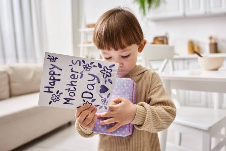 adorable lindo niño en cómoda ropa de casa posando con presente y tarjeta de felicitación en el día de las madres