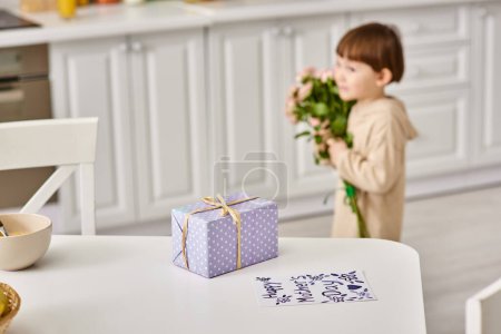 adorable niño en ropa de casa con ramo de flores cerca de la mesa con regalo y postal en ella