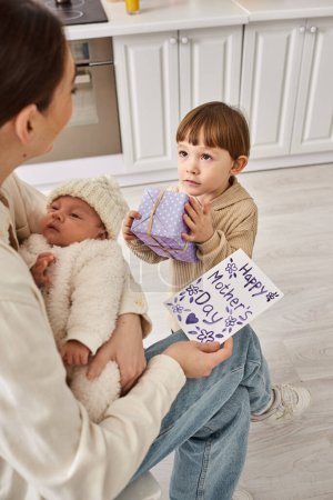 entzückender Junge, der seiner Mutter Geschenk und Postkarte gibt, während sie seinen neugeborenen Bruder hält, Muttertag