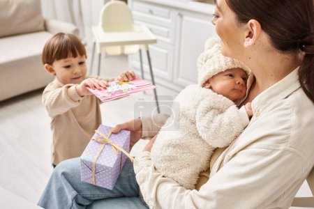 adorable boy giving present and postcard to mom while she holding his newborn brother, Mothers day