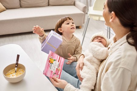 toddler boy giving present and postcard to mom while she holding his newborn brother, Mothers day