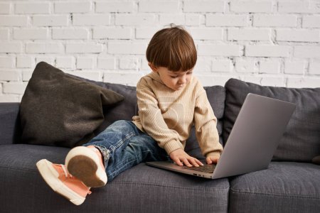 adorable lindo niño en acogedora ropa de casa sentado en el sofá y mirando el ordenador portátil con atención