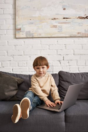 adorable lindo niño en acogedora ropa de casa sentado en el sofá con el ordenador portátil y mirando a la cámara