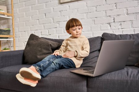 adorable lindo niño en acogedora ropa de casa sentado en el sofá y mirando a la computadora portátil con atención