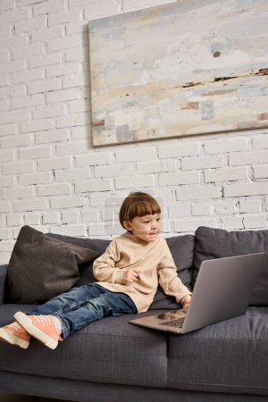 konzentrierte entzückende niedliche Kleinkind Junge in gemütlichen Homewear sitzt auf dem Sofa und schaut auf Laptop