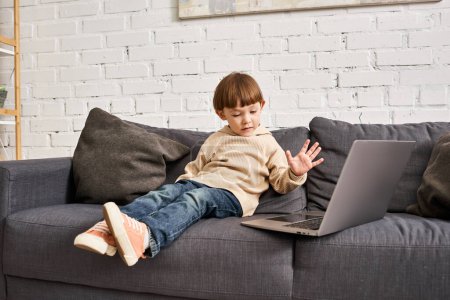 focused adorable cute toddler boy in comfortable homewear sitting on sofa and looking at laptop