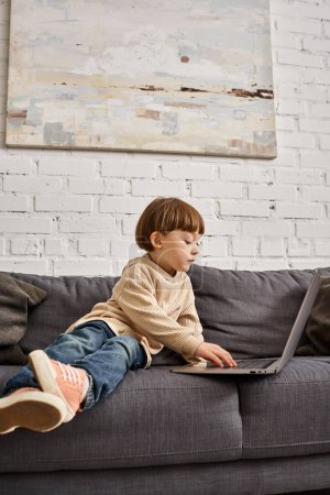 adorable cute toddler boy in casual homewear sitting on sofa and looking at laptop attentively