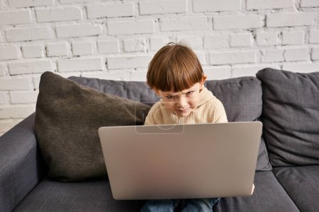 concentrado adorable lindo niño en acogedora ropa de casa sentado en el sofá y mirando el ordenador portátil