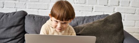 focused adorable cute toddler boy in cozy homewear sitting on couch and looking at laptop, banner