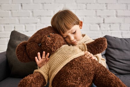 cute little boy in casual warm homewear hugging his teddy bear while sitting on couch at home