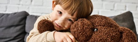 happy little boy hugging his teddy bear while sitting on couch and looking at camera, banner