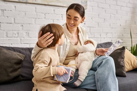 fröhliche hübsche Frau sitzt mit ihren Söhnen auf dem Sofa mit Geschenk und Grußkarte am Muttertag