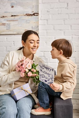 liebevolle lustige Mutter posiert auf dem Sofa mit ihrem kleinen Sohn mit Blumenstrauß und Geschenk, Muttertag