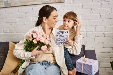 fürsorgliche fröhliche Mutter posiert auf dem Sofa mit ihrem kleinen Sohn mit Blumenstrauß und Geschenk, Muttertag