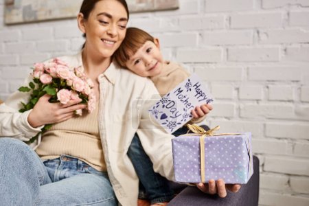 ziemlich glückliche Mutter posiert auf dem Sofa mit ihrem kleinen Sohn mit Blumenstrauß und Geschenk, Muttertag