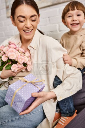 liebevolle glückliche Mutter posiert auf dem Sofa mit ihrem kleinen Sohn mit Blumenstrauß und Geschenk, Muttertag