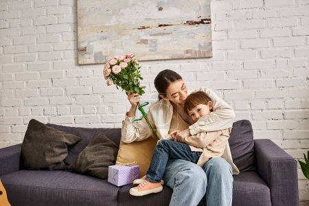 caring happy mother posing on sofa with her toddler son with flower bouquet and present, Mothers day