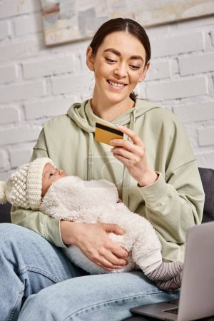 beautiful smiling mother in homewear holding her baby boy in font of laptop with credit card in hand
