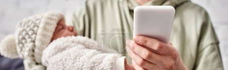 Photo for Loving mother in casual homewear holding her mobile phone while her newborn baby boy in her hands - Royalty Free Image