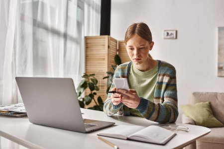 adolescente concentrada usando su teléfono inteligente mientras está sentado cerca de la computadora portátil en el escritorio, sesión de estudio electrónico