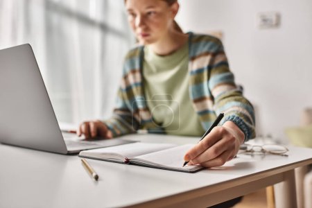 Konzentrierter Teenager macht Hausaufgaben am Laptop im häuslichen Umfeld, konzentriert sich darauf, dass das Mädchen Notizen macht