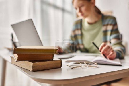 Photo for Focus on books on desk, teenage girl holding pen and looking at laptop while studying from home - Royalty Free Image