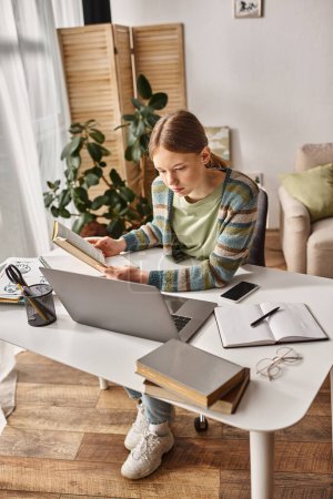 gen z girl holding book while using a laptop for e-learning at home, teen lifestyle concept