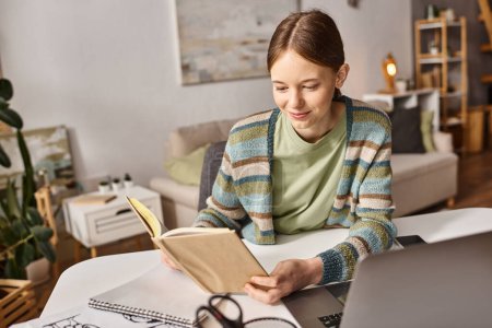 Teenager Mädchen, das Buch in der Hand hält, während es zu Hause einen Laptop zum E-Learning benutzt, Lebensstil der Generation Z