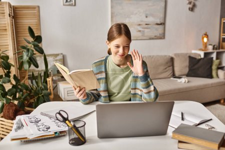 happy gen z girl holding book while using a laptop during video call at home, teen lifestyle concept