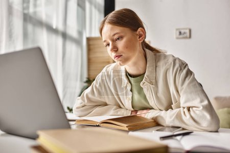 Focused teenager doing homework on laptop in a home environment, focus on gen z girl near books