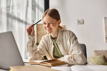 Téléchargez les photos : Adolescent positif faisant des devoirs sur ordinateur portable dans un environnement à la maison, se concentrer sur la fille gen z près des livres - en image libre de droit